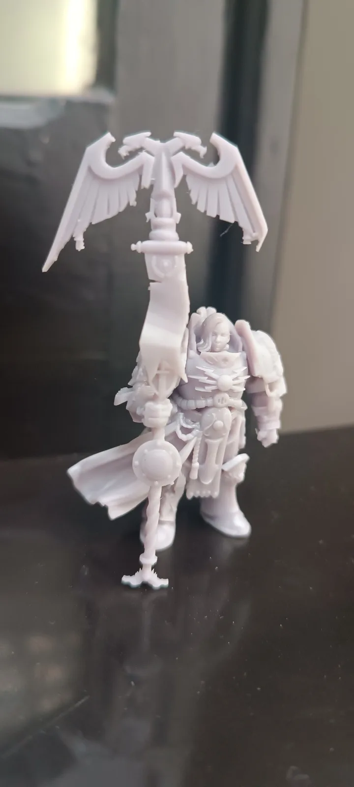 A white, unpainted miniature figurine of a heavily armored warrior holding a tall staff topped with a large, spread-winged…