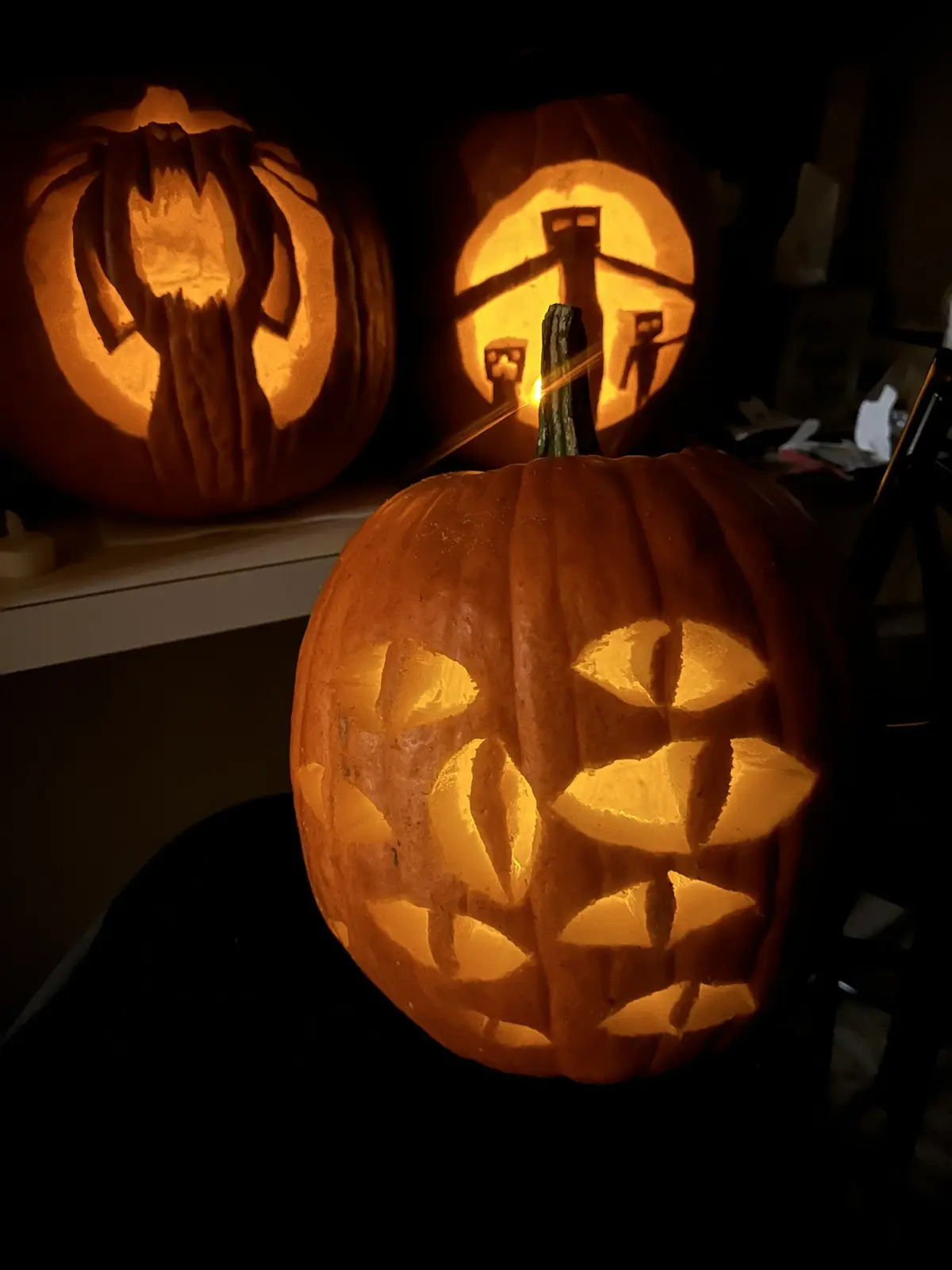 Three carved pumpkins glow from within in a dark room, with the foreground pumpkin featuring a grid of cut-out eyes and…