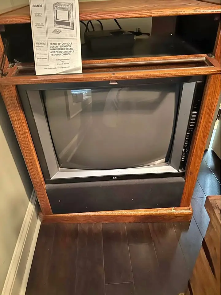 A vintage CRT television in a wooden console cabinet, with a Sears owner's manual visibly propped up on the shelf above it…