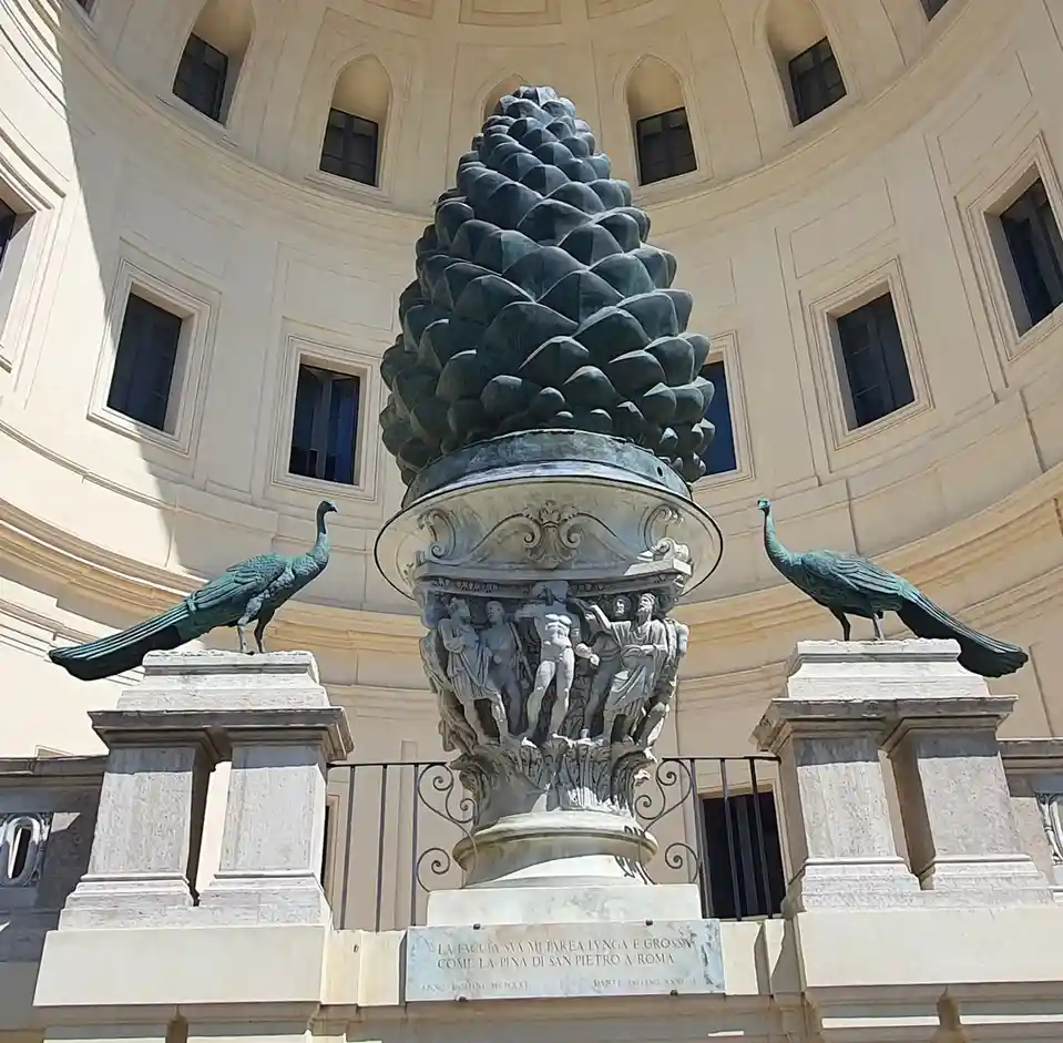 A large bronze pinecone sculpture sits atop a stone pedestal in a circular building, flanked by two bronze peacocks on…