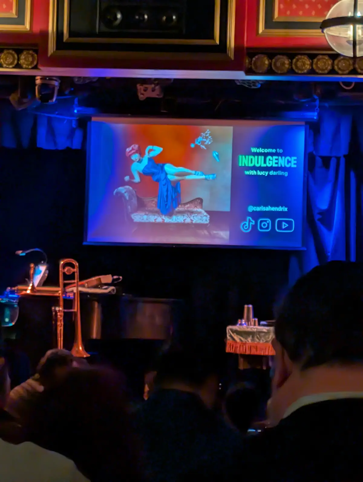 A dimly lit venue with audience members watching a screen displaying a vintage-style illustration of a woman in a blue dress…