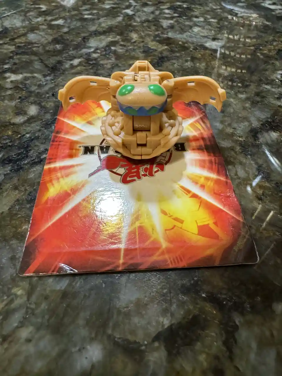 A tan plastic Beyblade toy with green eyes and bat-like wings sits atop its red and orange Beyblade card, which has visible…