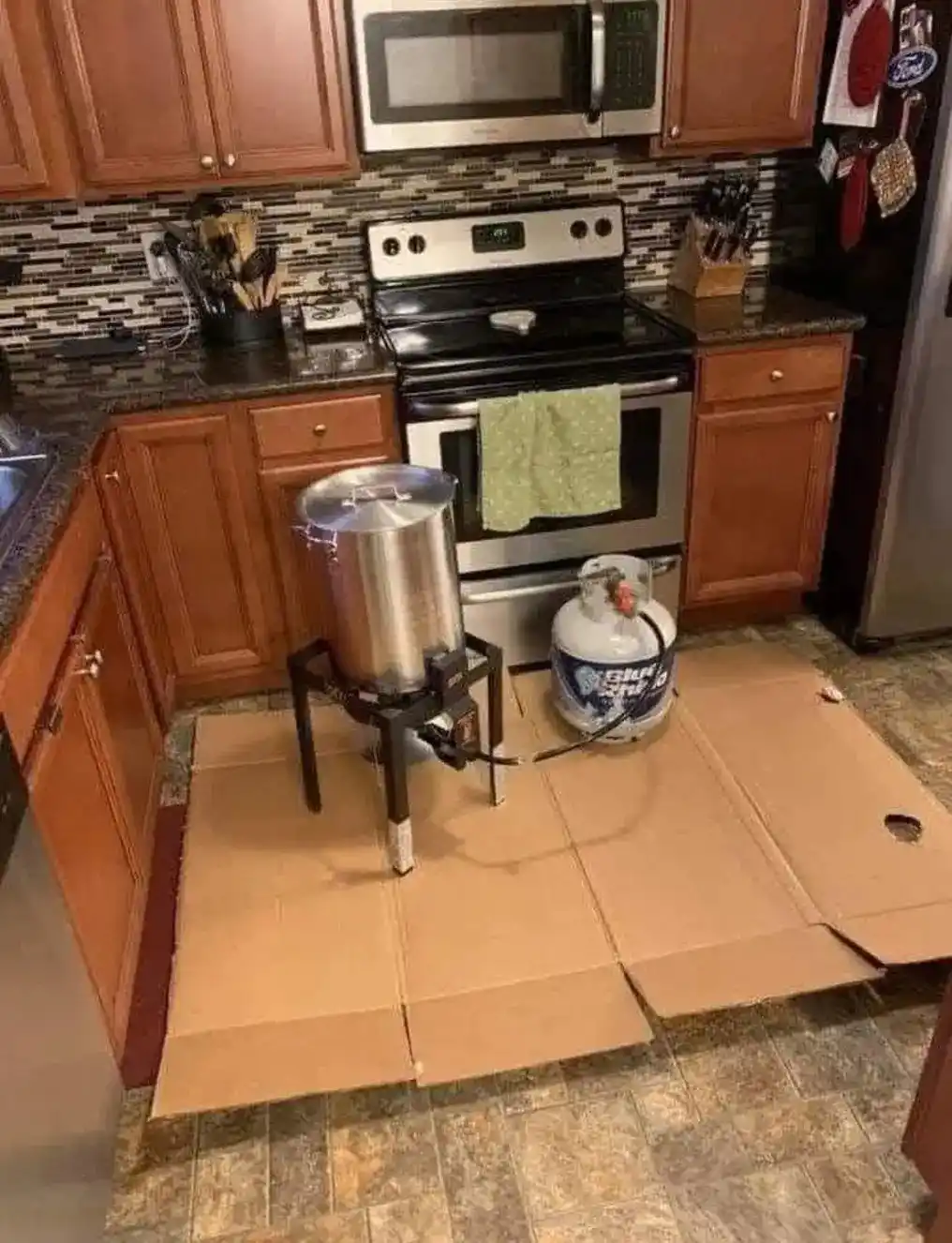 A kitchen scene shows a large metal pot on a portable burner connected to a propane tank, set on a makeshift cardboard floor…