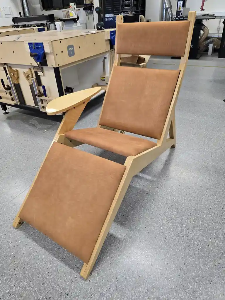 A wooden recliner chair with tan leather cushions sits in a workshop, surrounded by woodworking tools and workbenches. The…