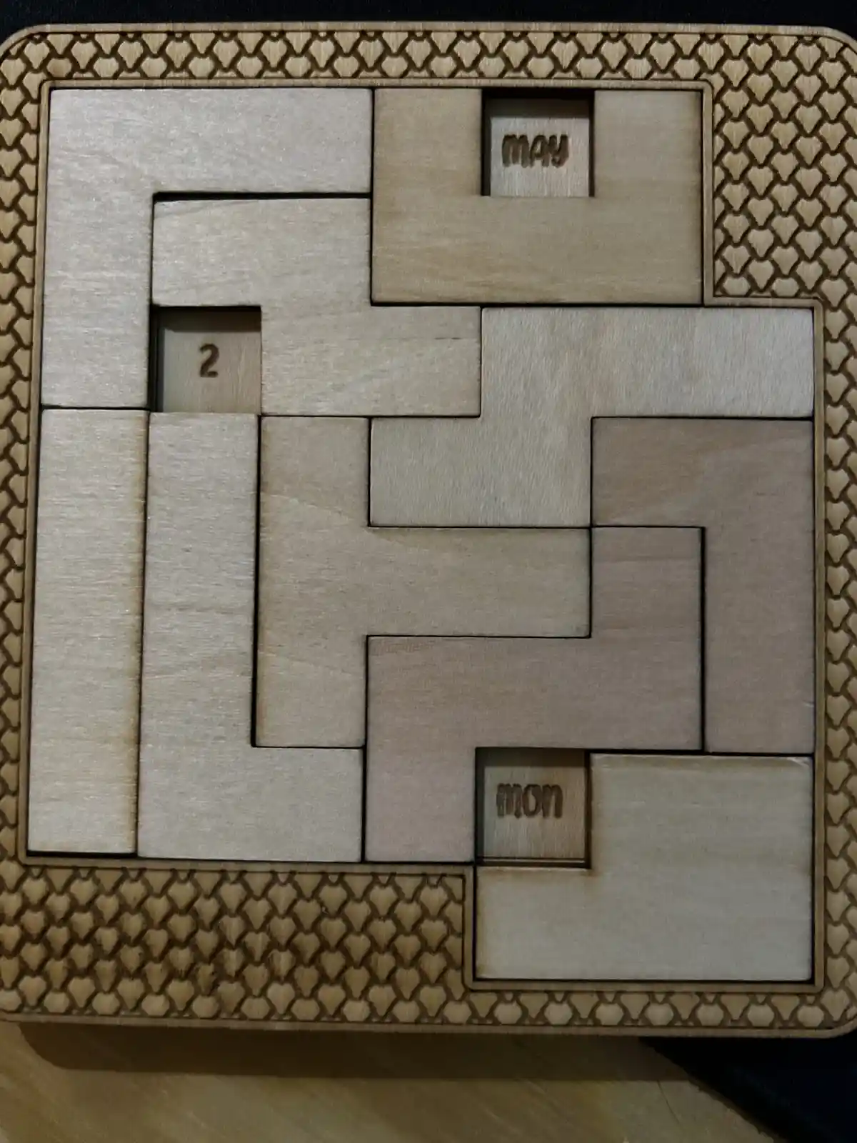 A wooden puzzle with interlocking pieces forms a maze-like structure, featuring two small blocks labeled 'may' and 'mon' in…
