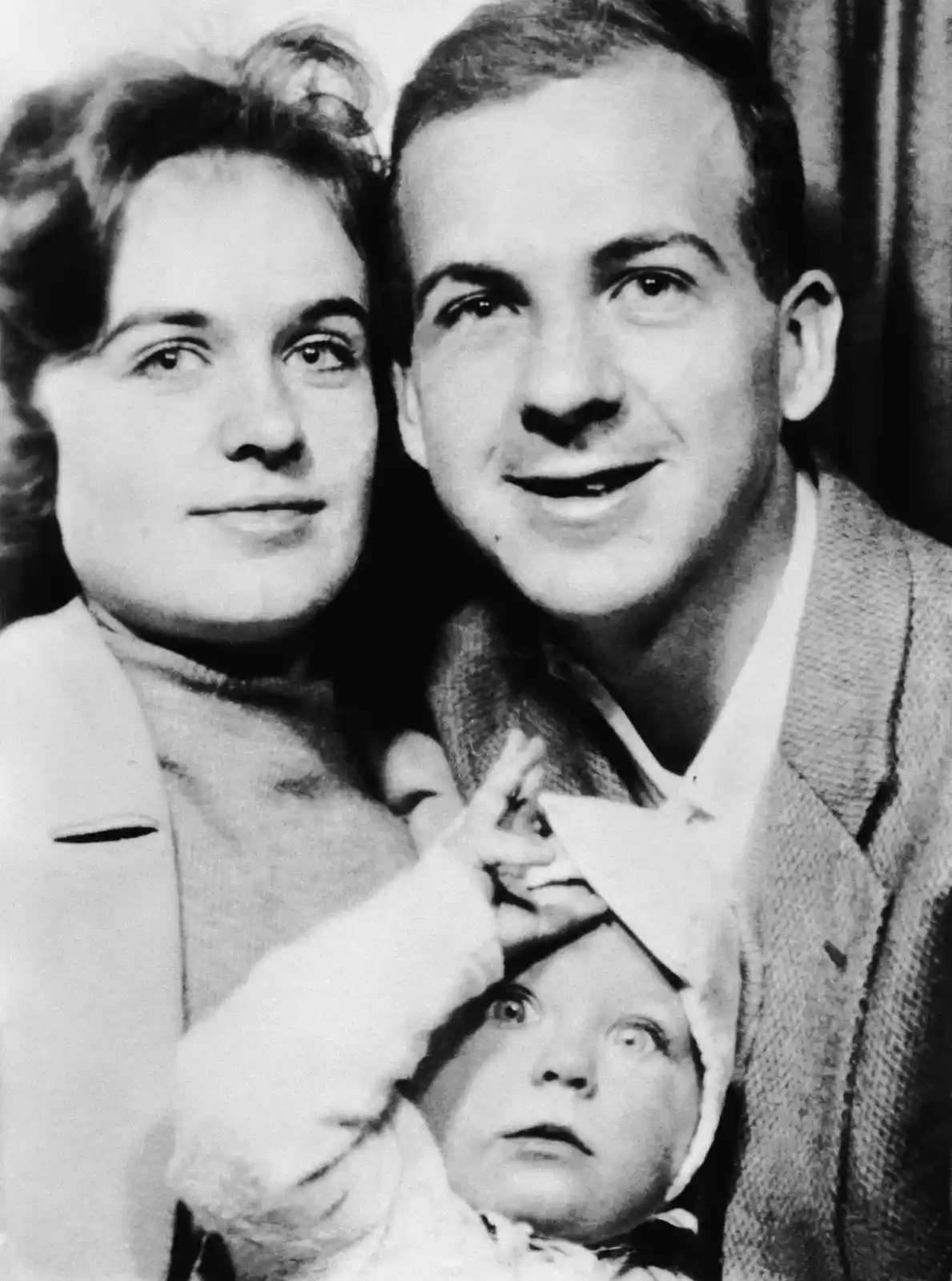 A vintage black-and-white family photo shows a man and woman posing with a baby. The baby, wearing a bonnet, has wide…