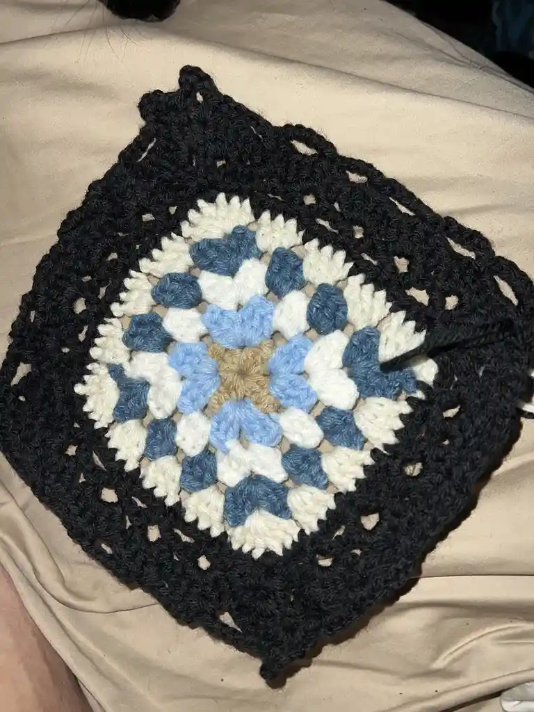 A hand-crocheted circular doily with a black outer border, white and blue floral patterns, and a beige center rests on a…