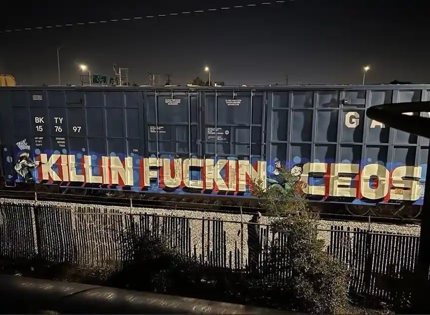 A nighttime photo of a dark blue freight train car with graffiti that reads 'KILLIN FUCKIN CEOS' in large, stylized letters…