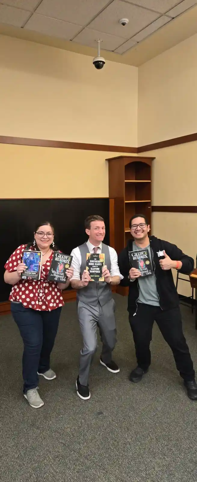 Three people stand in a room holding books by Grady Hendrix, smiling and posing for the camera. The person in the center…