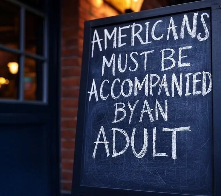 A close-up of a black chalkboard sign with white chalk text reading 'AMERICANS MUST BE ACCOMPANIED BY AN ADULT', placed…