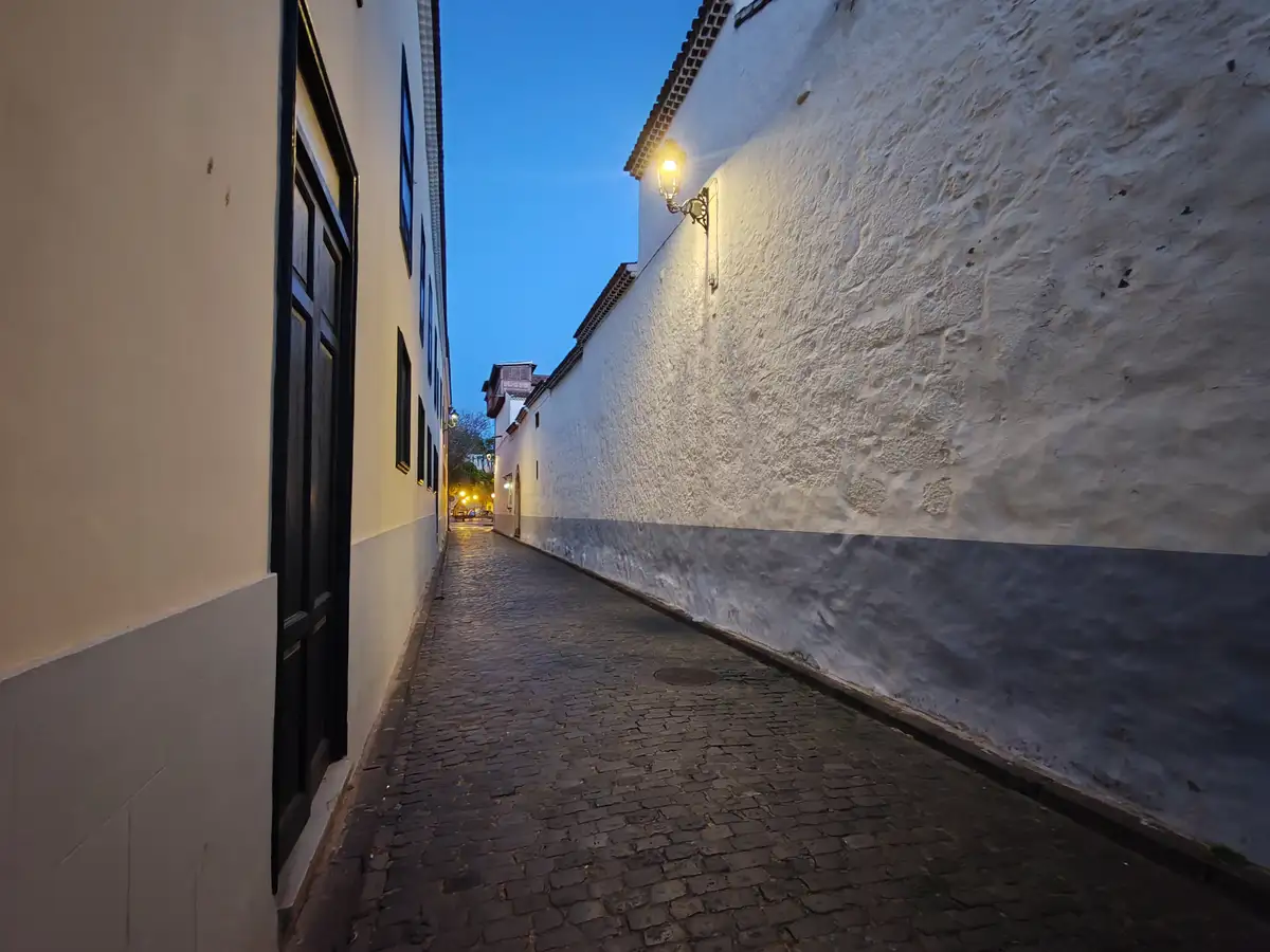 A narrow cobblestone alleyway at dusk, flanked by white stucco walls with a single lit wall lamp casting warm light. The…