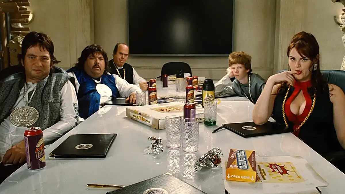 A group of people, including a woman in a red top and gold chain, sit around a conference table with pizza boxes, soda cans…