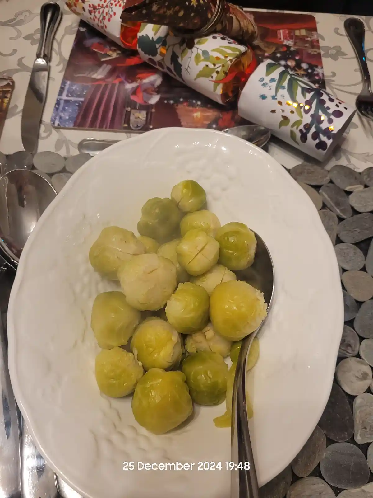 A white plate holds boiled Brussels sprouts with a spoon resting in them, set on a festive table with Christmas crackers and…