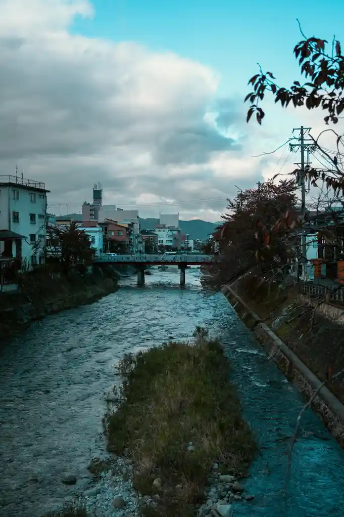 A serene, slightly moody photograph of a river flowing through a town with buildings on both sides and a bridge in the…