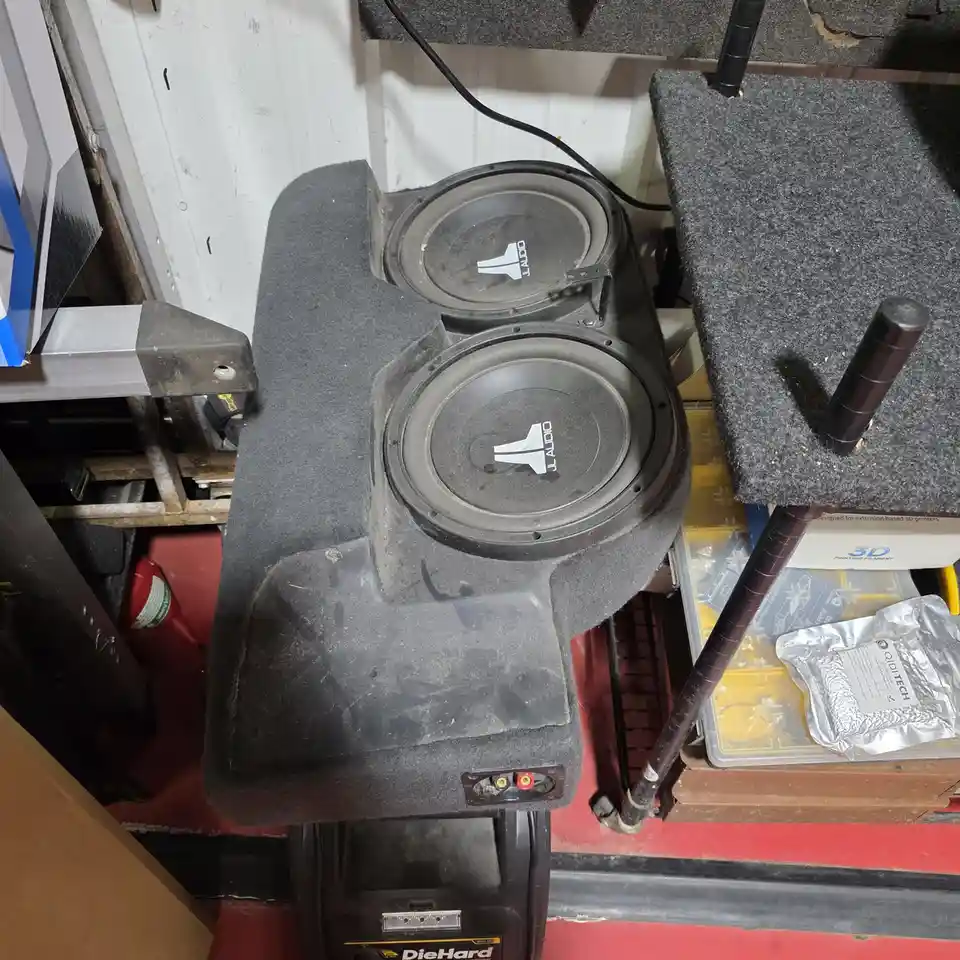 A dusty, custom-built car subwoofer enclosure with two JL Audio subwoofers sits on a workbench in a cluttered garage…