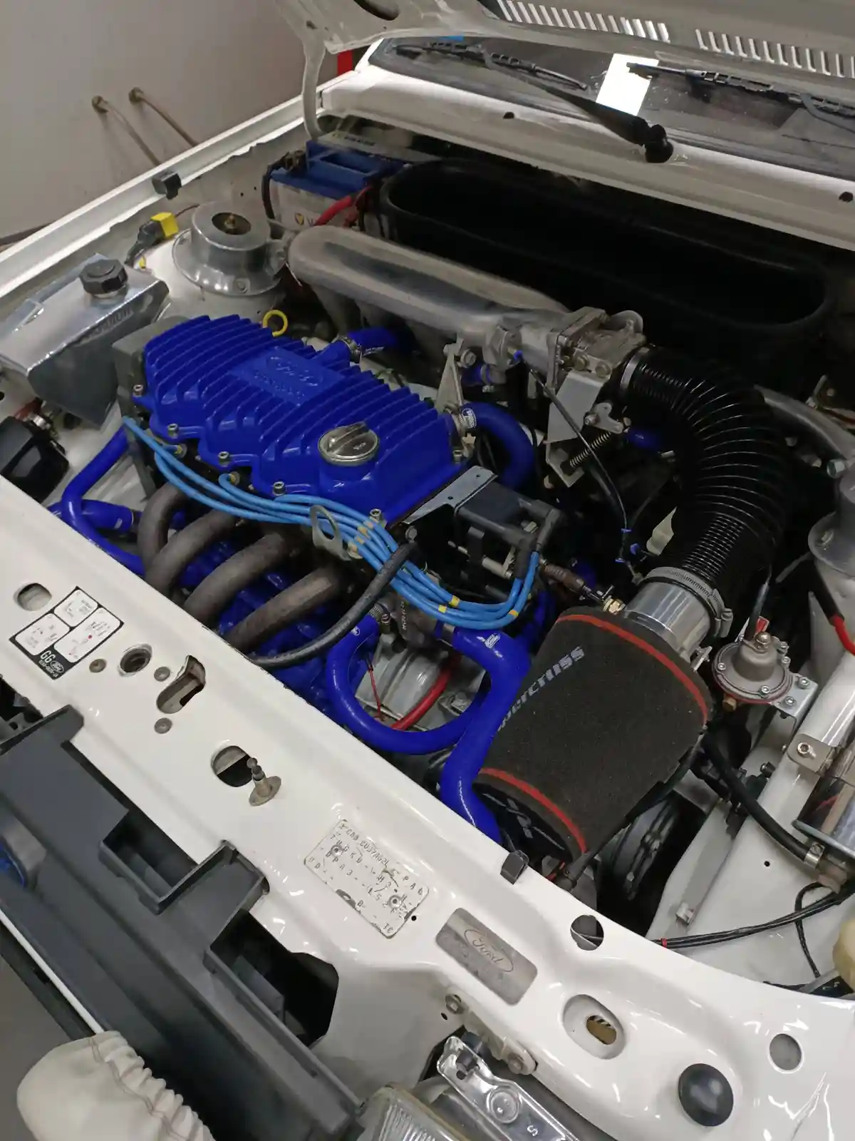 A close-up photo of a white car's engine bay with a prominently featured blue aftermarket engine cover, blue hoses, and a…