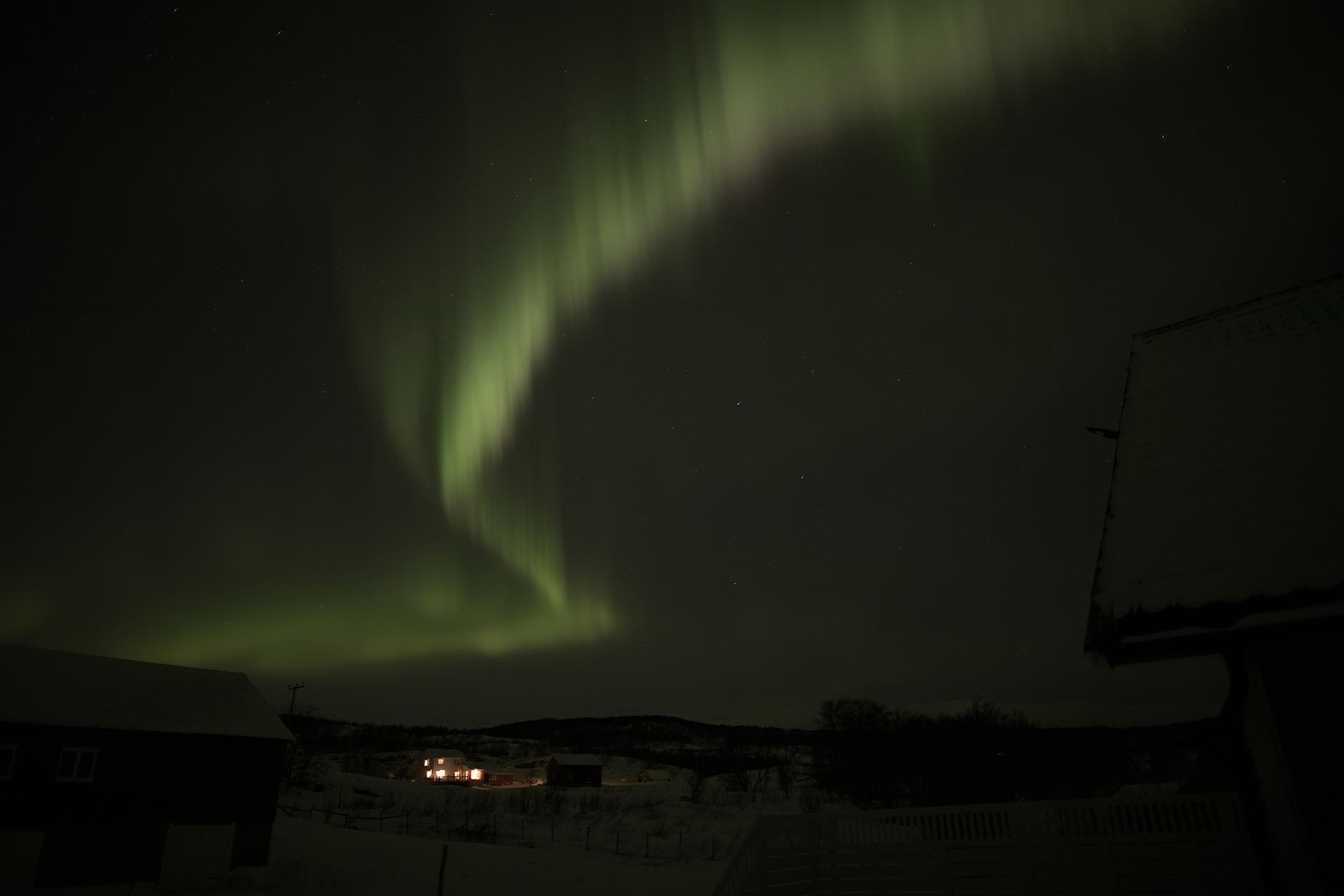 A nighttime photograph shows a vibrant green aurora borealis dancing across a starry sky above a snowy landscape with…