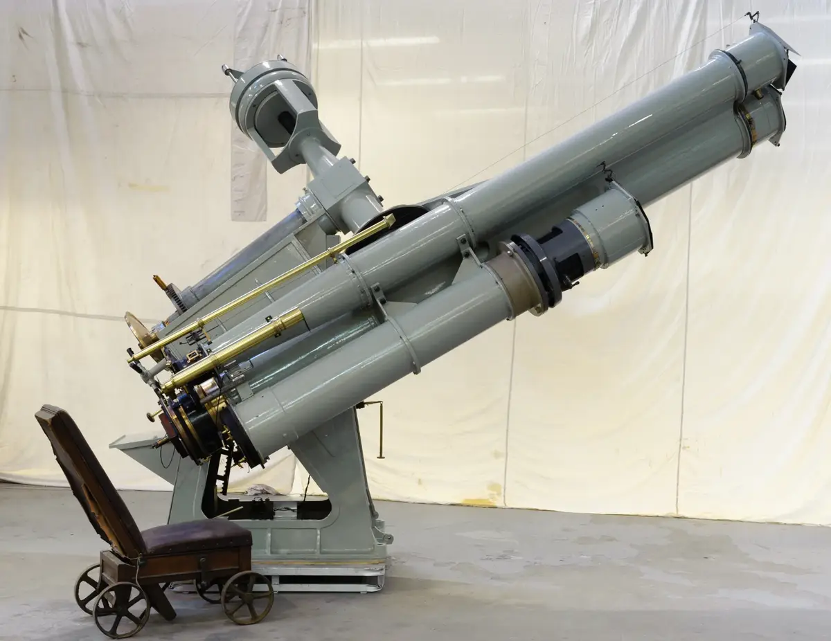 A large, grey, vintage-style astronomical telescope mounted on a heavy base, angled upward. Beside it is a small, wheeled…