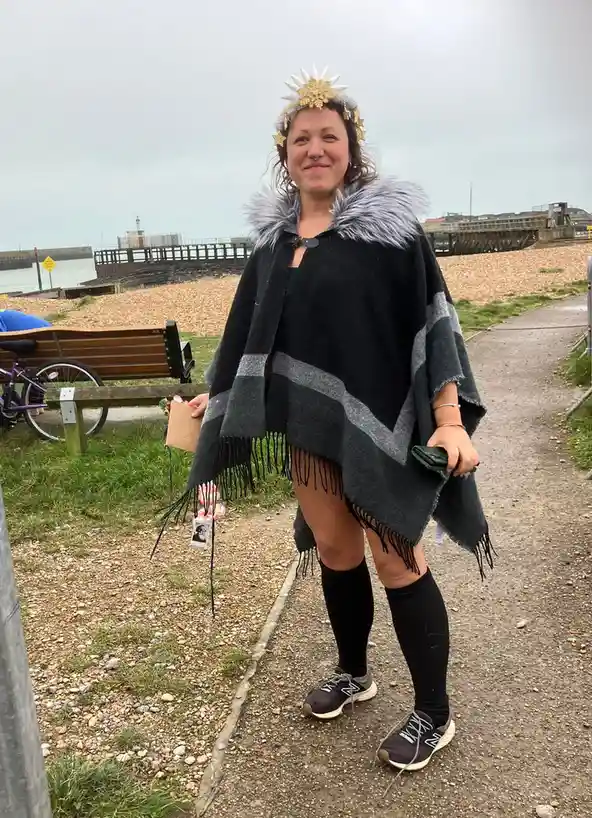 A woman stands outdoors on a gravel path near a beach, wearing a black and grey striped poncho with a silver faux fur…
