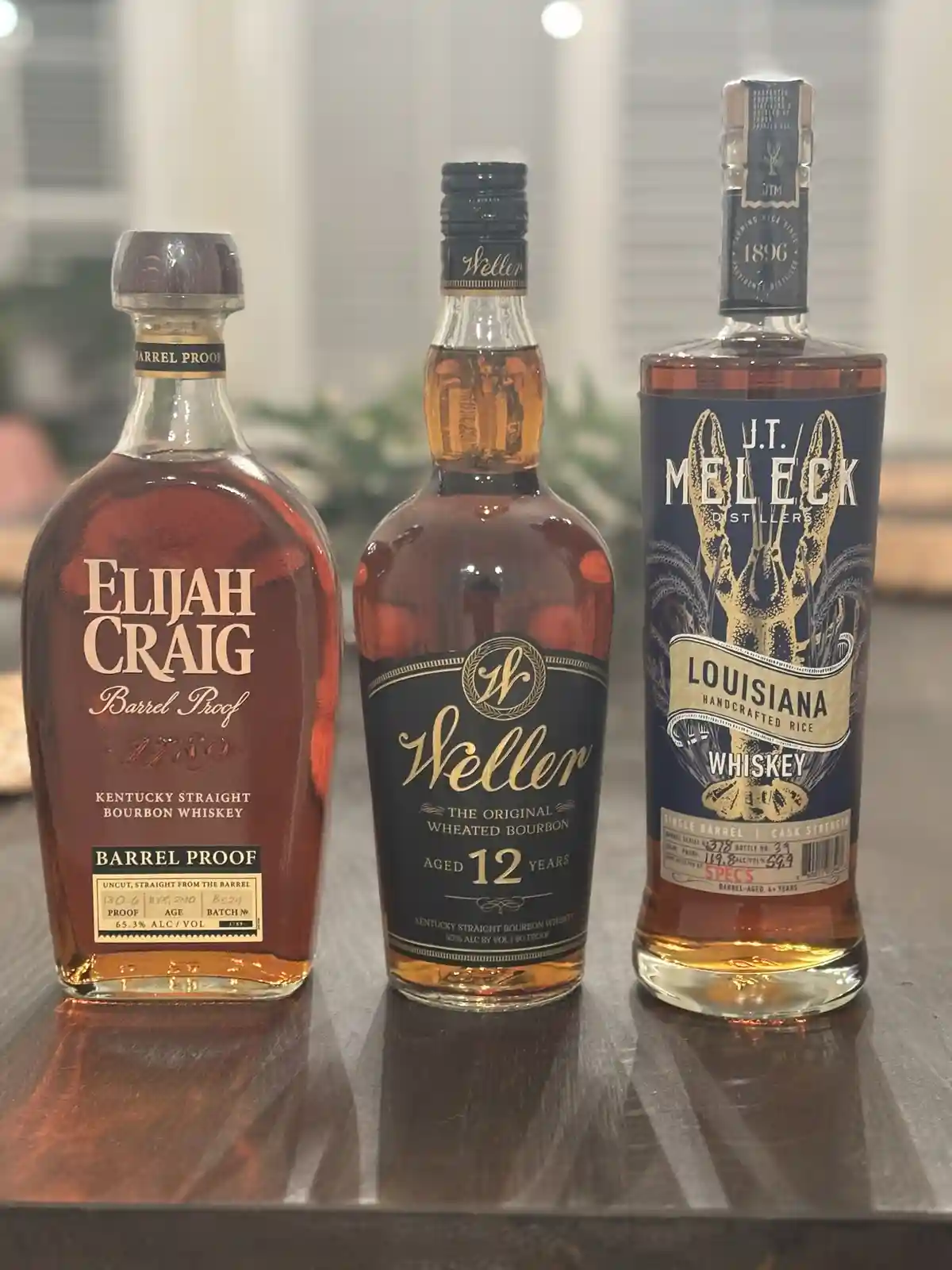 Three bottles of whiskey are lined up on a dark countertop: Elijah Craig Barrel Proof, Weller 12-Year-Old, and J.T. Meleck…