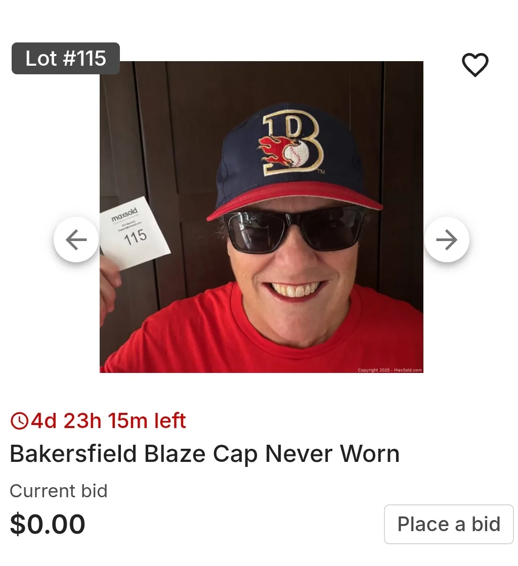 A man wearing a blue and red baseball cap with a 'B' logo and sunglasses smiles while holding up a 'maxsold' auction tag…