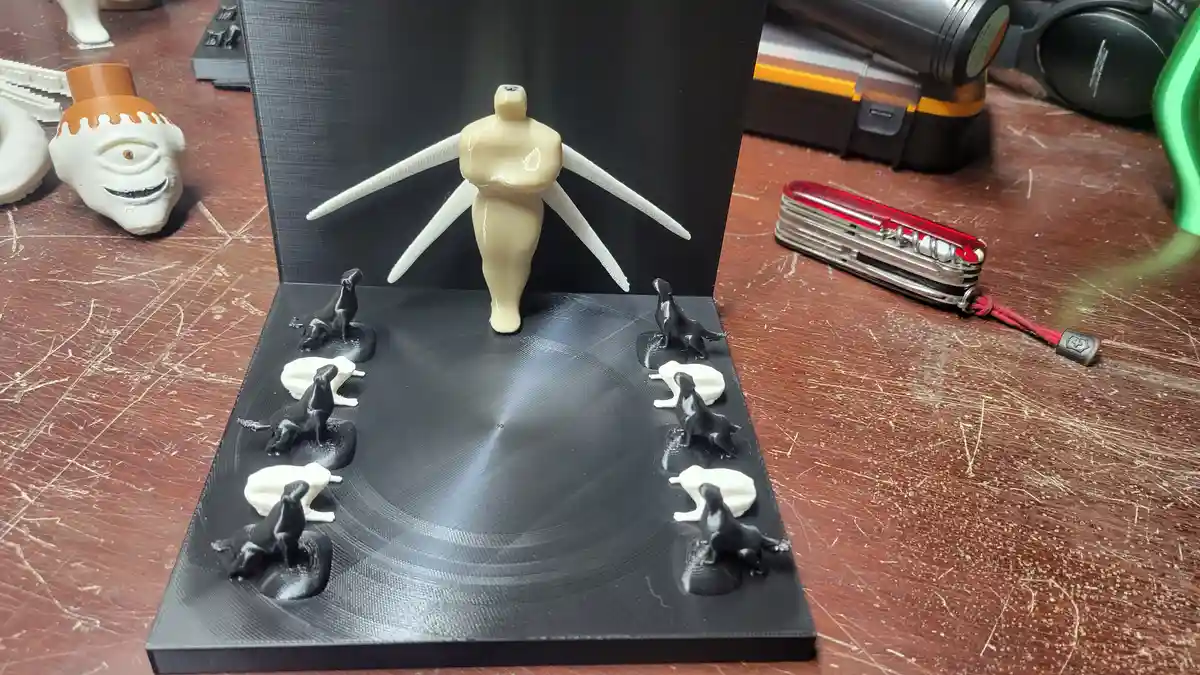 A 3d-printed figurine of a nude female goddess with white wing-like structures stands on a black circular platform…