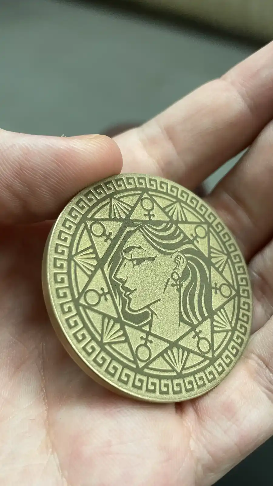 A hand holds a brass-colored coin featuring a stylized profile of a woman with flowing hair, surrounded by geometric…