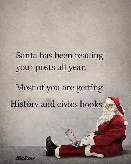 A man dressed as Santa Claus sits cross-legged on a gray floor against a textured gray wall, using a silver laptop. Black…