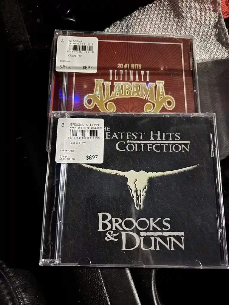 Two country music CD cases are stacked, both priced at $6.97. The top case is 'Ultimate Alabama' with a red cover and gold…