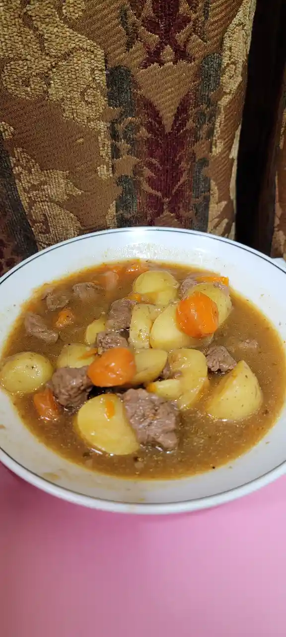 A white bowl with a blue rim contains a hearty stew of chunks of meat, potatoes, and carrots in a brown broth, placed on a…