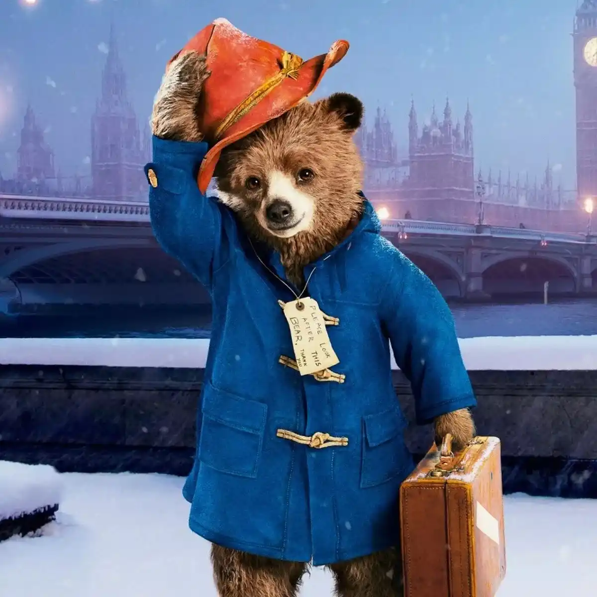 A brown bear wearing a blue duffle coat and red hat stands in a snowy London scene with Big Ben and a bridge in the…