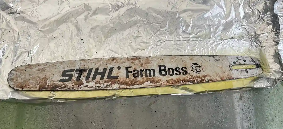 A heavily rusted and dirty STIHL Farm Boss chainsaw bar lies on crumpled aluminum foil, with visible wear and dirt covering…