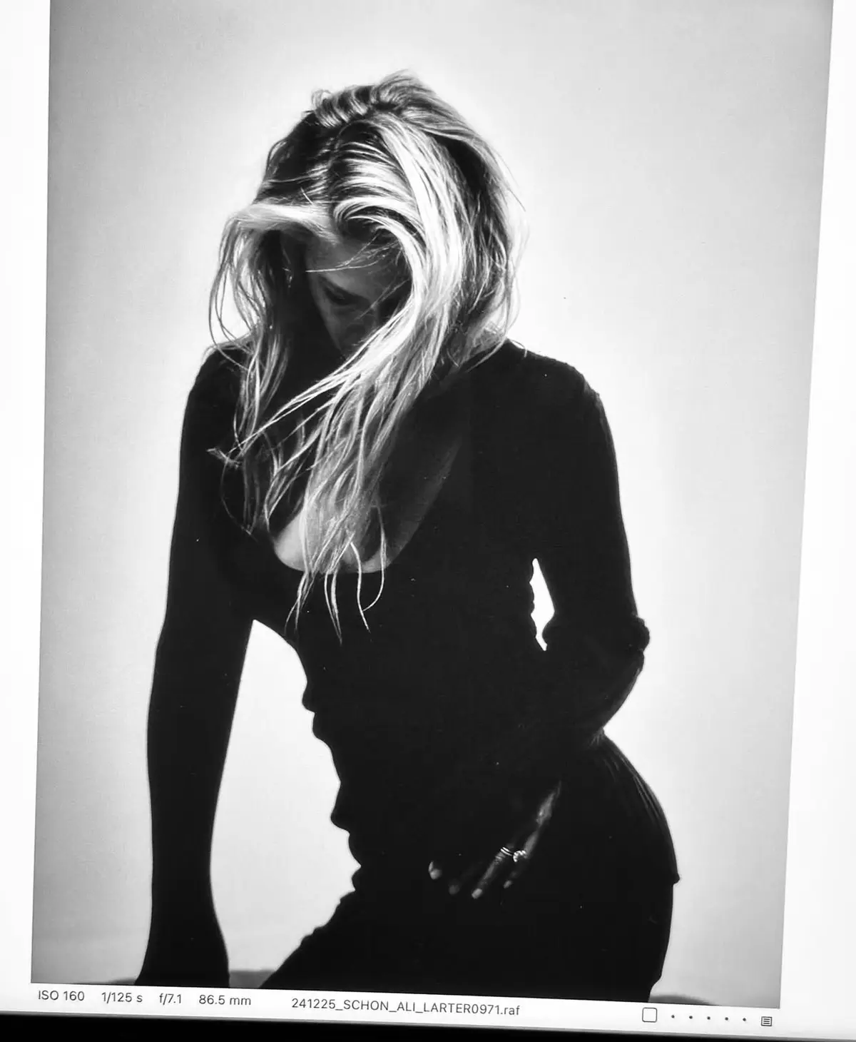 A black and white photo shows a woman with long blonde hair covering her face as she leans forward, wearing a dark top. The…