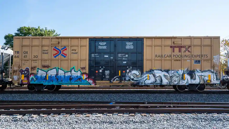 A tan freight railcar with 'TTX RAILCAR POOLING EXPERTS' branding and graffiti art sits on tracks under a clear blue sky…