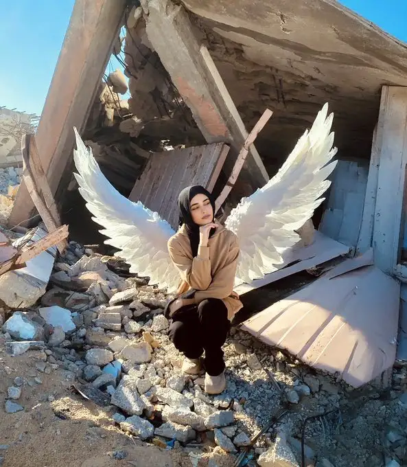 A woman wearing a hijab and large white angel wings sits amidst rubble and debris from a collapsed building. She looks…