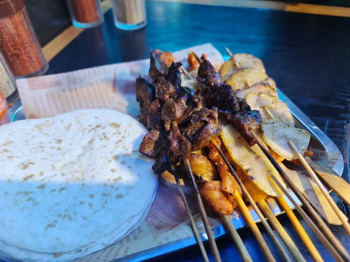 A close-up photo of a metal tray holding multiple skewers of grilled meat and vegetables alongside a stack of flatbread…