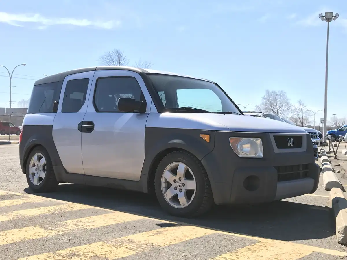 A silver and black Honda Element is parked in a lot under a clear blue sky. The car has a boxy, rugged design with visible…