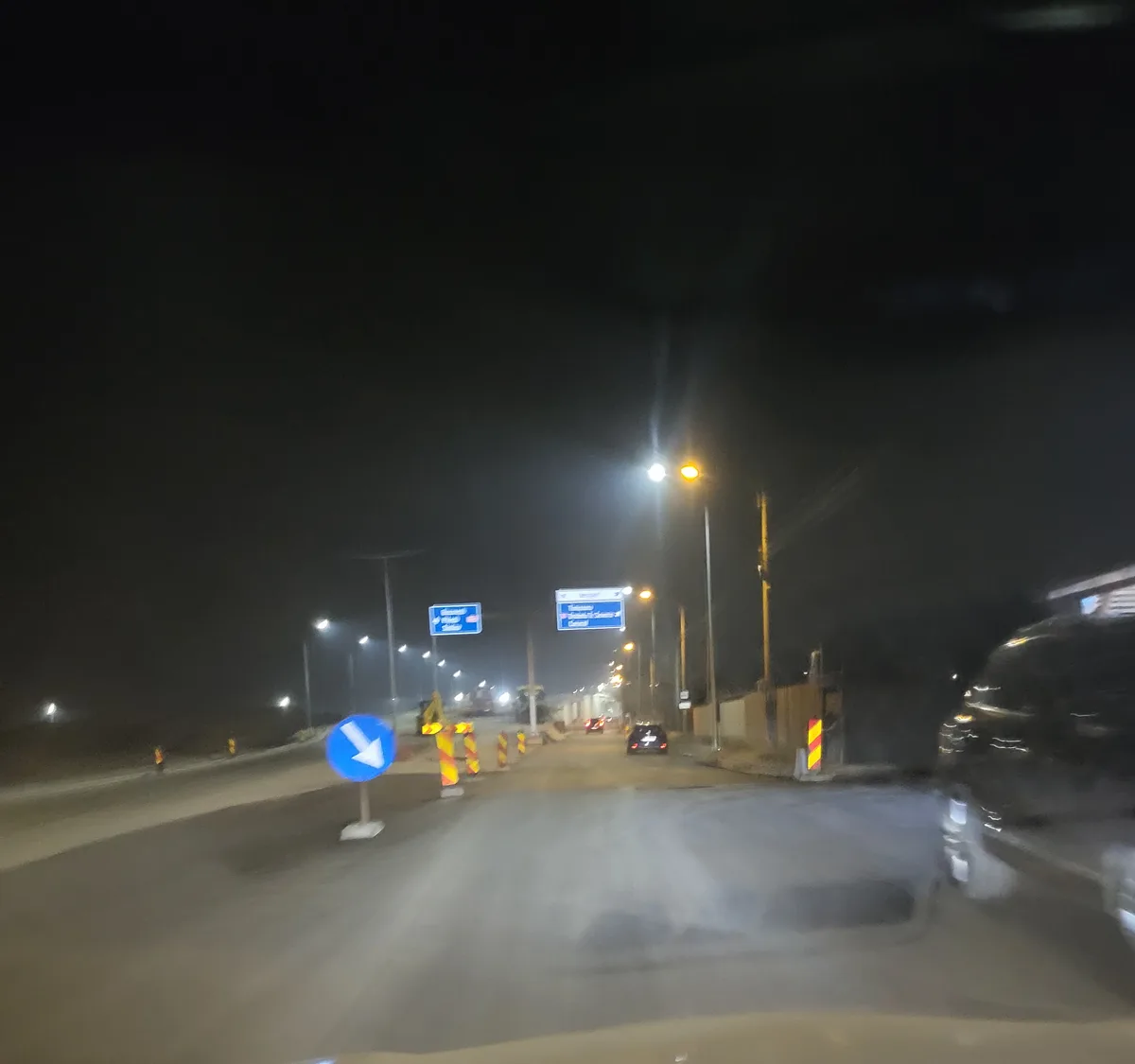 A nighttime dashcam view from a car driving on a road with construction, showing a blue circular sign with a white arrow…