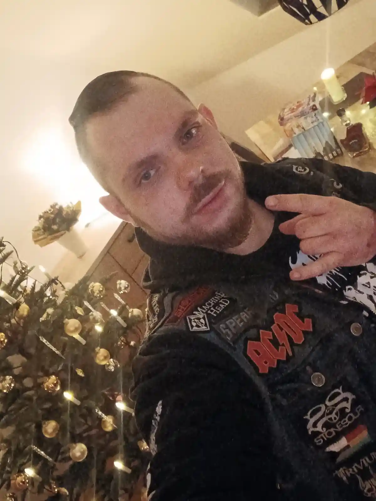 A man with a mustache and short hair takes a selfie in front of a decorated christmas tree with gold ornaments and lights…