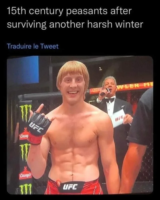 A shirtless, muscular man with a wide, triumphant grin and a blonde mullet stands in a UFC octagon, wearing red shorts and…