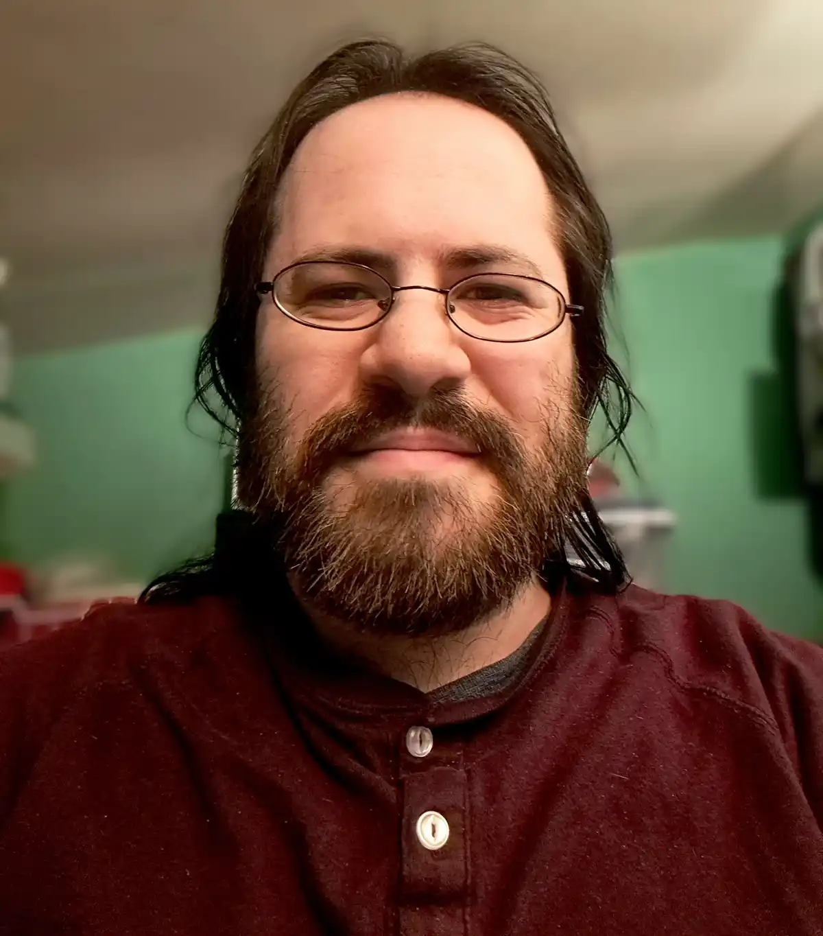A man with long dark hair, glasses, and a beard wearing a maroon button-up shirt smiles slightly while looking at the…