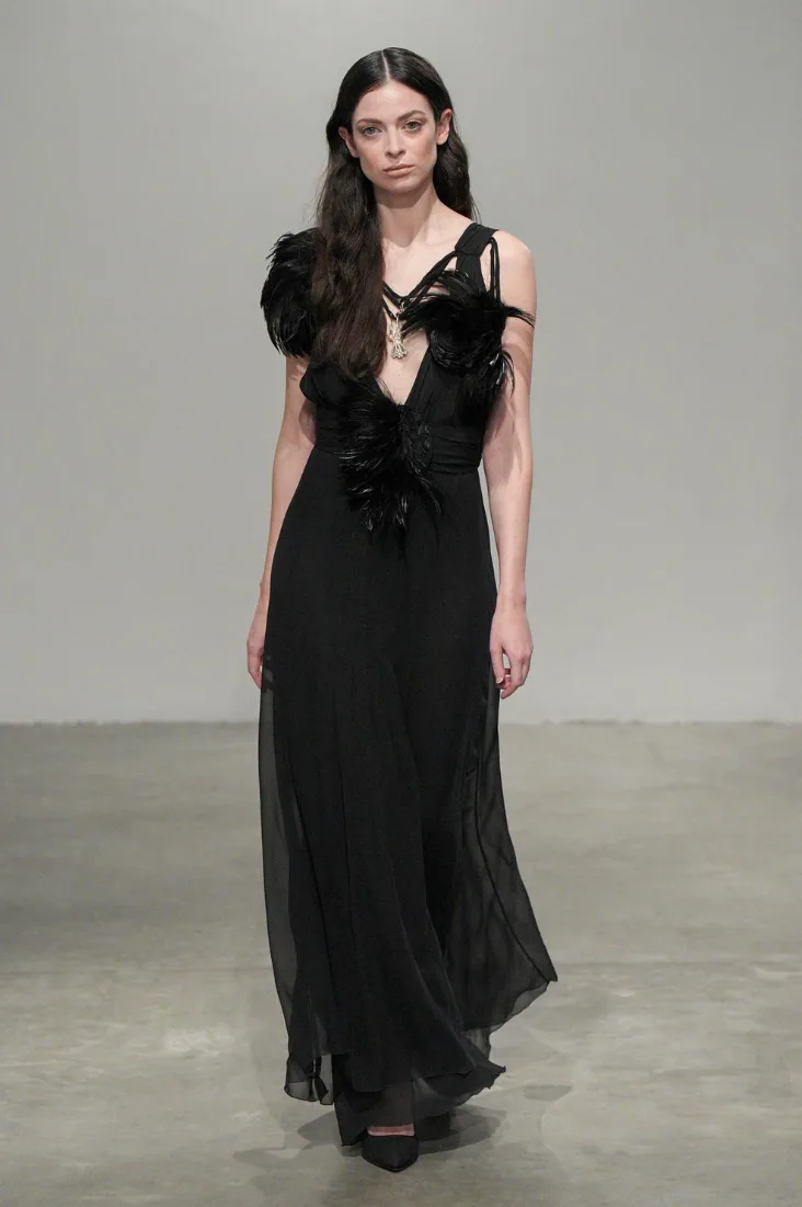 A model walks down a runway in a black, sheer, floor-length gown with feathered shoulder and chest embellishments. She has…
