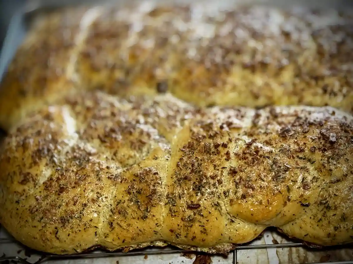 A close-up photo of a golden-brown, herb-topped braided bread cooling on a wire rack. The bread has a glossy surface with…