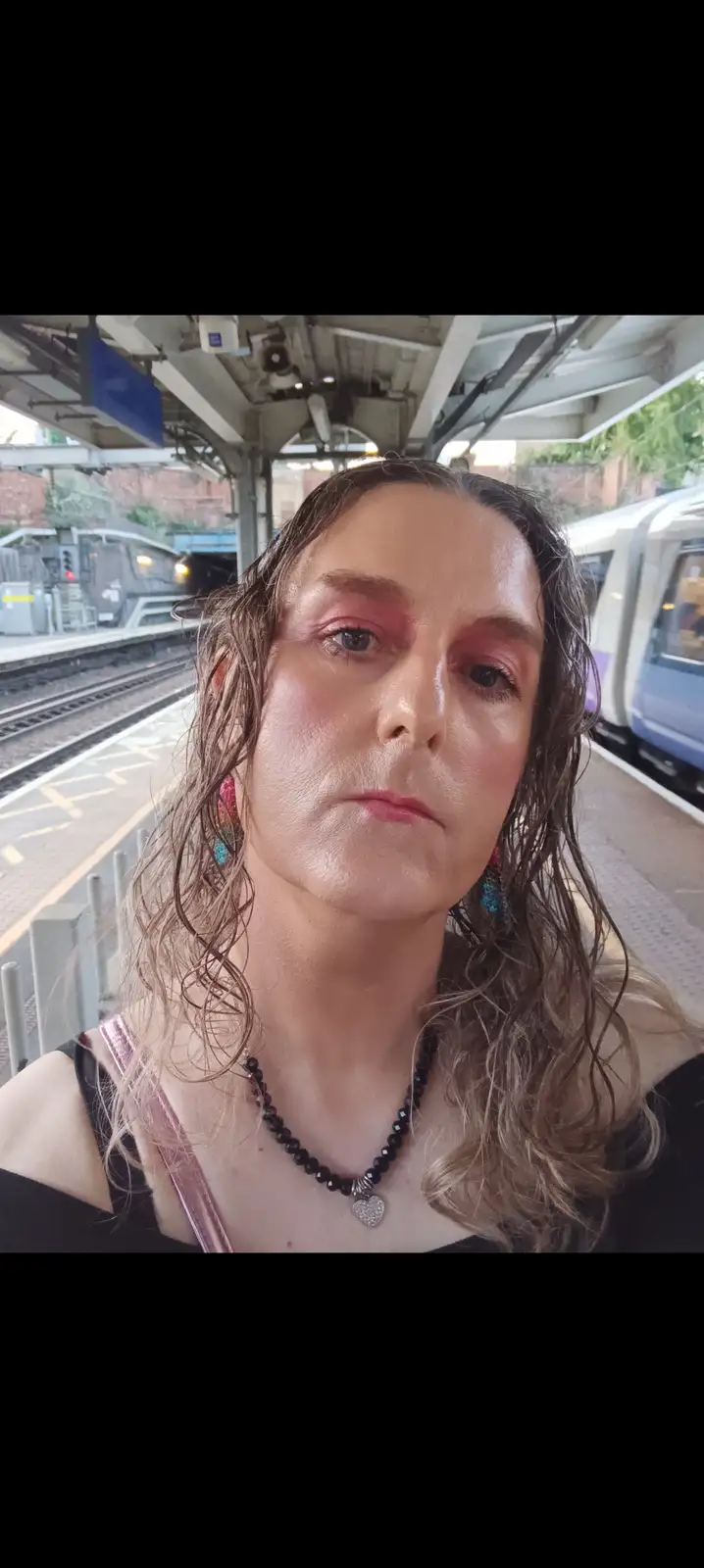 A person with wet, messy hair and smudged pink eye makeup stands on a train platform, looking directly at the camera with a…