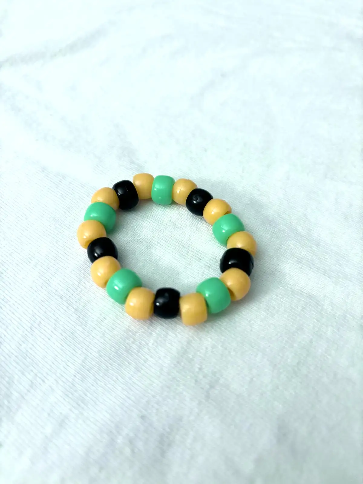 A close-up photo shows a handmade beaded bracelet with alternating green, yellow, and black beads resting on a white…