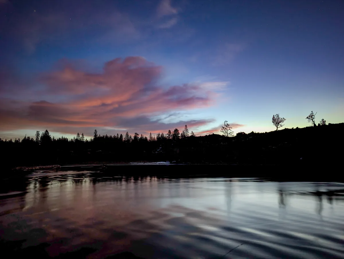 A serene twilight landscape featuring a calm river reflecting a starry sky with pink and purple clouds, silhouetted trees…