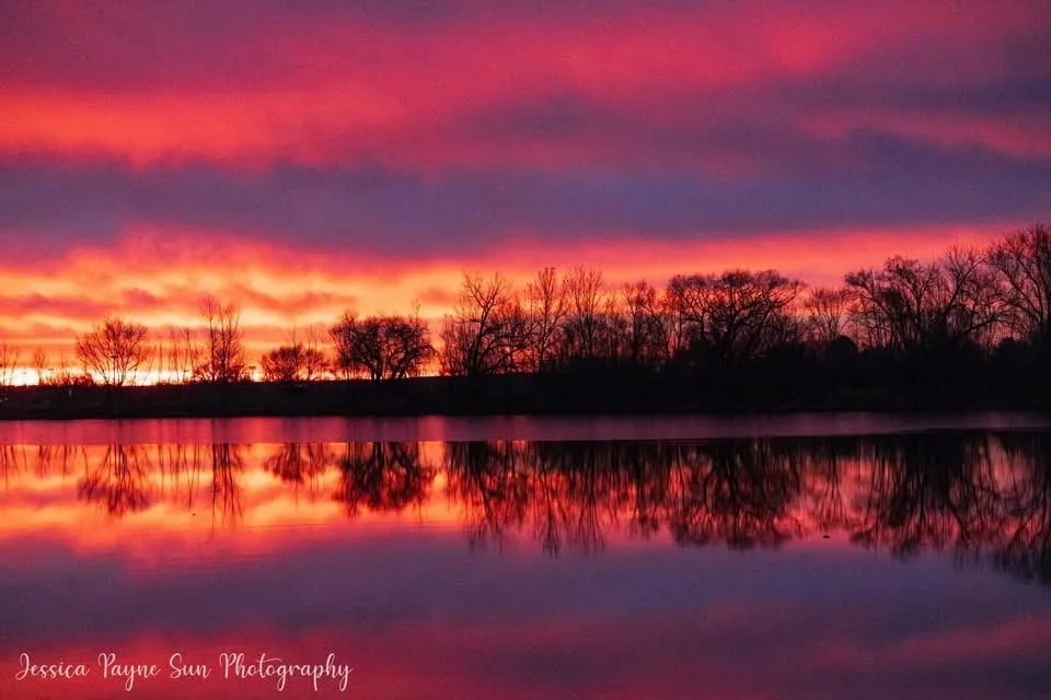 A serene landscape photograph shows a vibrant sunset with fiery orange, pink, and purple clouds reflected perfectly in a…