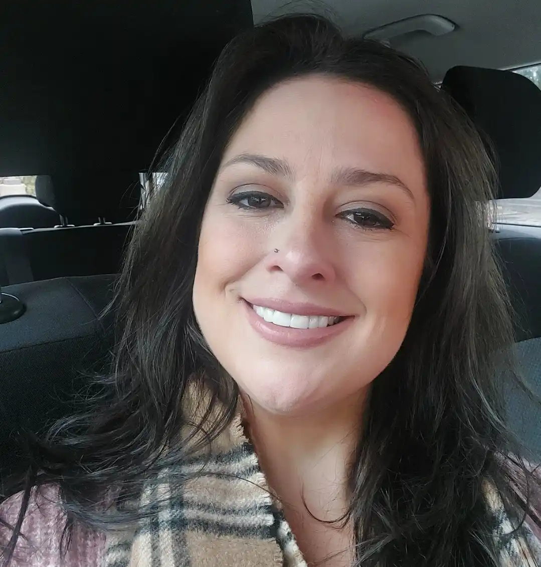 A woman with dark hair and a nose piercing smiles broadly while sitting in a car, wearing a plaid scarf. The overlaid text…