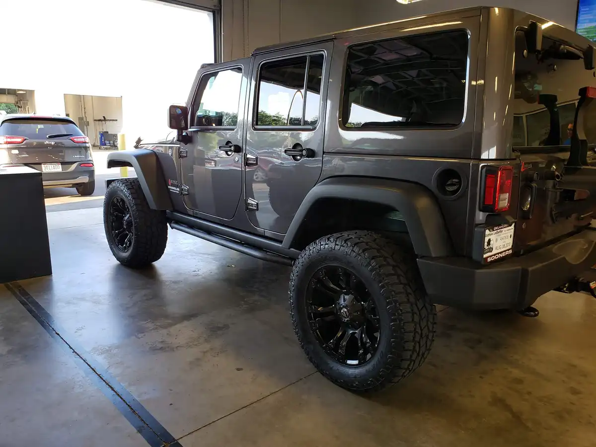 A dark gray Jeep Wrangler Unlimited is parked inside a garage, featuring black wheels and rugged tires. Another Jeep is…