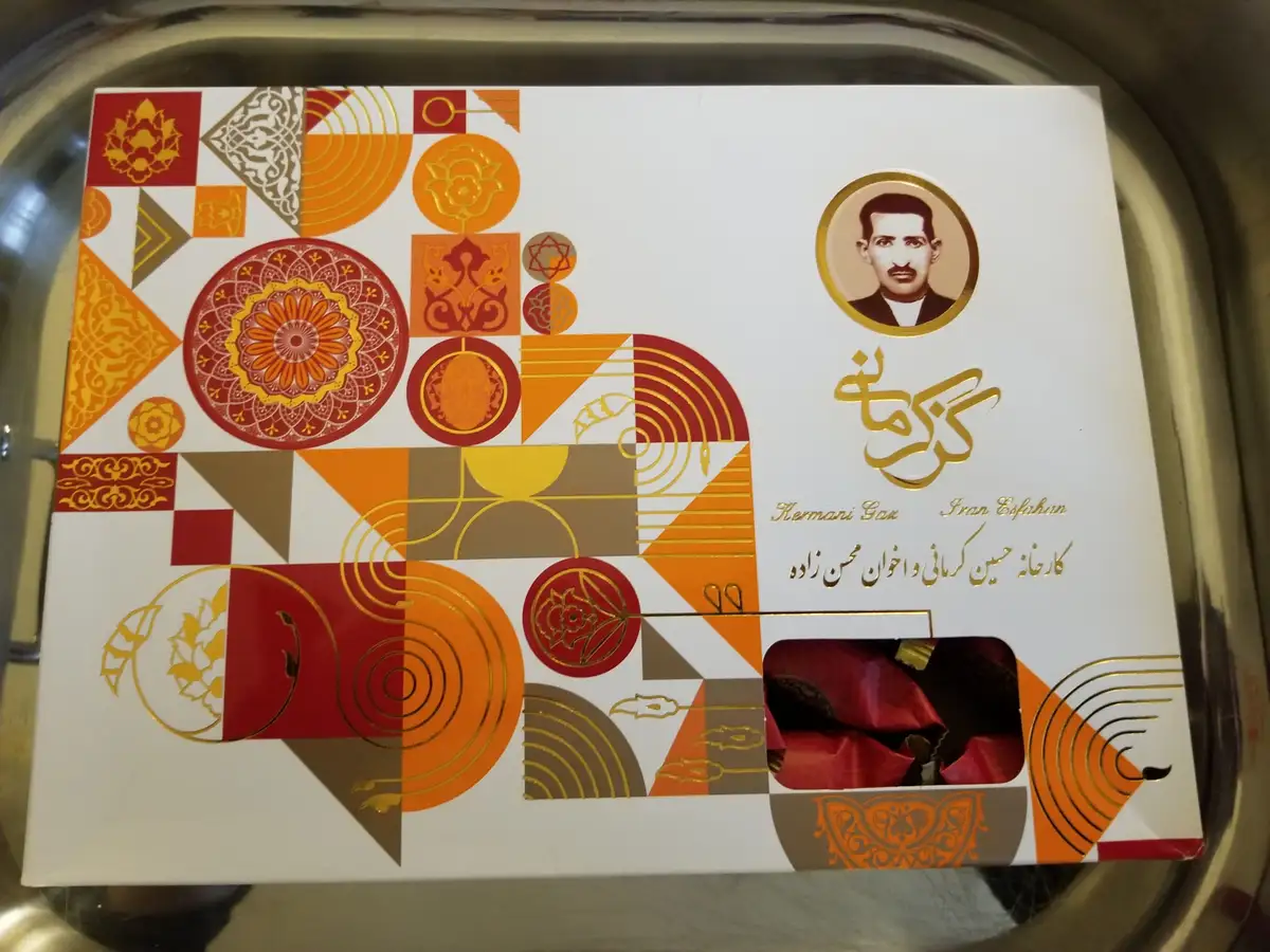A decorative white box with red, orange, and gold geometric and floral patterns sits on a metal tray. It features a circular…